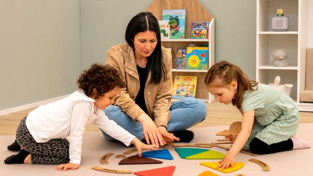 Erzieherin und 2 Kindern sitzen auf dem Boden im Morgenkreis und spielen mit einem Wochenkreis aus Holz