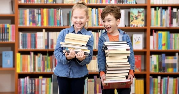Zwei Kinder stehen in einer Bibliothek und tragen jeweils einen Stapel Bücher 