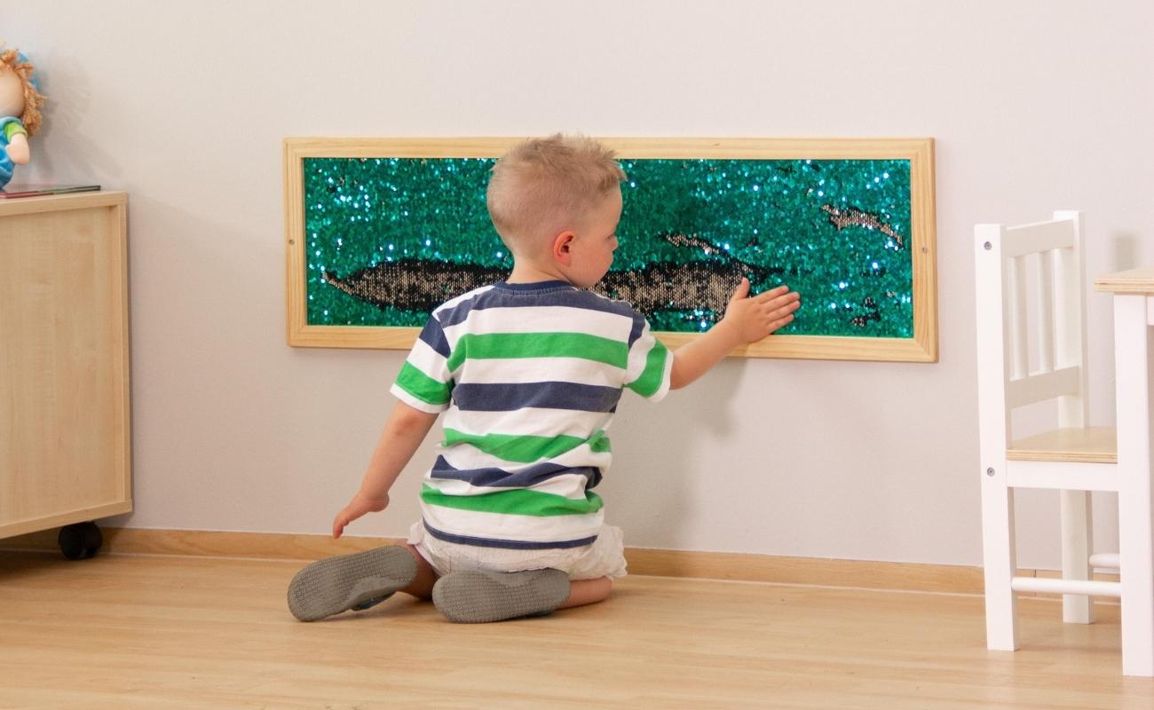 Junge sitzt auf dem Boden und spielt mit Pailletten-Wandboard